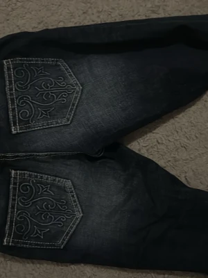 Mörkblå bootcut jeans med brodyr - Snygga mörkblå bootcut jeans med vita kontrastsömmar och stora bakfickor med dekorativ brodyr. Jeansen har en klassisk femficksmodell och lätt slitna detaljer framtill. Perfekta för dig som vill ha en unik touch på din denimstil.