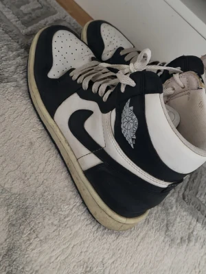 Svarta och vita Air Jordan 1 sneakers - Säljer ett par klassiska Air Jordan 1 sneakers i svart och vitt läder med vita snören och ikonisk vinge-logga på sidan. Skorna har perforerad tåbox och svart swoosh. Perfekta för dig som gillar streetwear och tidlös design.