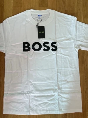 Vit BOSS t-shirt med tryck - Vit t-shirt från BOSS med klassisk rund hals och svart BOSS-logga tryckt framtill. T-shirten har korta ärmar och är tillverkad i mjuk bomull, perfekt för en clean och stilren look. Pris går att diskutera.