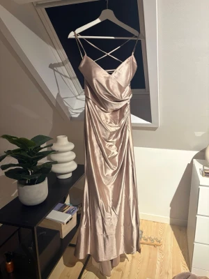 Champagnefärgad långklänning i satin - Säljer en elegant långklänning i champagnefärgad satin med tunna axelband och snygg snörning i ryggen. Klänningen har draperad front och en smickrande siluett som ger en lyxig känsla. Perfekt för dig som vill sticka ut på nästa event!