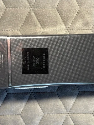 Tom Ford Oud Wood Eau de Parfum - Tom Ford Oud Wood Eau de Parfum i en elegant, mörkgrå förpackning med diskret mönster och svart etikett. Flaskan rymmer 100 ml och doften är känd för sina exklusiva tränoter och lyxiga känsla. Perfekt för dig som gillar unika och sofistikerade parfymer.