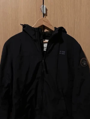 Marinblå vindjacka från Napapijri - Snygg vindjacka från Napapijri med huva och dragkedja framtill. Jackan har en patch med logga på ärmen och diskret flaggdetalj på bröstet. Perfekt för dig som vill ha en stilren och funktionell jacka.