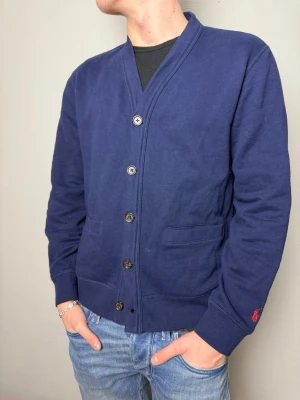 Marinblå cardigan från Polo Ralph Lauren - Stilren marinblå cardigan från Polo Ralph Lauren med knäppning framtill och två fickor. Tröjan har ribbade muddar, förstärkta armbågar och en liten röd logga broderad på ärmen. Funkar perfekt både casual och lite formellt 