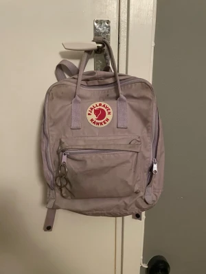 Ljuslila Kånken ryggsäck från Fjällräven - Säljer en ljuslila Fjällräven Kånken ryggsäck med klassisk fyrkantig form och det ikoniska röda rävmärket framtill. Ryggsäcken har ett stort huvudfack, en framficka med dragkedja och två sidofickor. Tillverkad i slitstarkt syntetmaterial och har justerbara axelband.