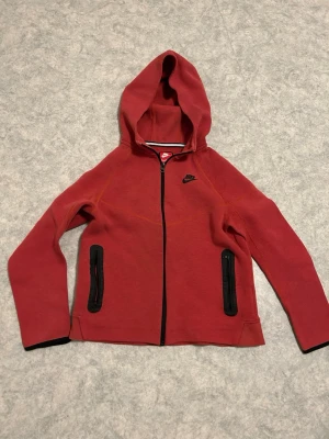 Röd hoodie från Nike med dragkedja - Röd hoodie från Nike med svart logga på bröstet och hel dragkedja framtill. Två svarta dragkedjeförsedda fickor på sidorna och huva. Tillverkad i mjukt och bekvämt material, perfekt för en avslappnad stil.