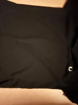 Svart piké från Fred Perry - Klassisk svart pikétröja från Fred Perry med vit broderad logga på bröstet. Tröjan har korta ärmar, krage och knappar framtill. Perfekt för en stilren och sportig look.