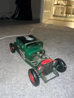 Retro Hot Rod i metall – vintage samlarobjekt / inredning - Säljer en retroinspirerad leksaksbil i metall med grön kaross, röda hjul och detaljer i silver. Bilen har en hajmun-dekal på sidan och ett flygplansmärke på taket. Motorn är detaljerad och fronten har röd grill. Perfekt för dig som gillar samlarprylar eller cool inredning.