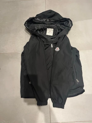 Svart vindjacka från Moncler - Snygg svart vindjacka från Moncler med huva och dragkedja framtill. Jackan har Moncler-logga på bröstet och tryckt Moncler-text på insidan av huvan. Två sidofickor med dragkedja och elastiska muddar vid ärmsluten. Perfekt för blåsiga dagar.väldigt fin jacka 