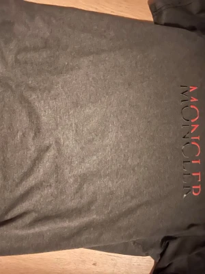 Grå Moncler t-shirt med tryck - Snygg grå t-shirt från Moncler med logotyp i svart och rött tryck längs sidan. Klassisk rund halsringning och korta ärmar. Perfekt för en stilren och avslappnad look.