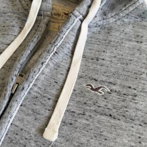Hollister Hoodie S - Snygg gråmelerad hoodie från Hollister med dragkedja framtill, vita snören i huvan och broderad logga på bröstet. Mjuk bomullsblandning och två fickor framtill. Perfekt för en avslappnad och trendig stil. Fraktar inom 1-2 dagar 📦