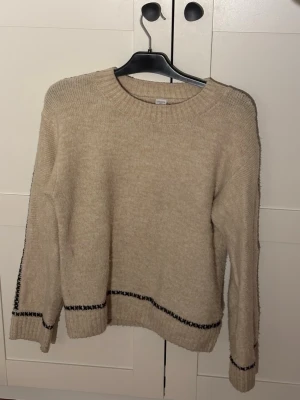 Beige stickad tröja med svart detalj - Mysig beige stickad tröja med rund hals och svarta korsstygnsdetaljer vid ärmslut och nederkant. Tröjan har en avslappnad passform och långa ärmar. Perfekt för kalla dagar och enkel att matcha med olika outfits.