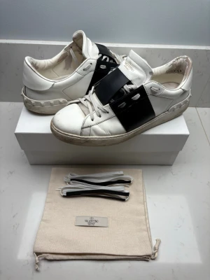 Valentino Garavani vita sneakers - Kom gärna med prisförslag! Snygga vita sneakers från Valentino Garavani med en bred svart läderdetalj över mitten och ikoniska nitar på hälen. Skorna har klassisk låg profil och kommer med extra skosnören och dustbag. Perfekta för dig som gillar exklusiv streetstyle. Org pris 7000kr+!