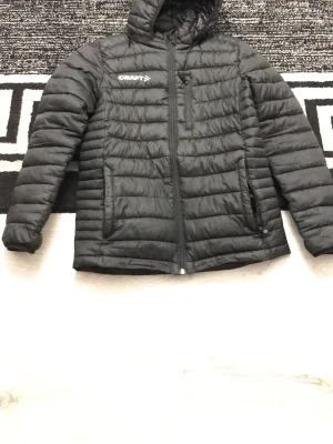 Svart pufferjacka från Craft - Säljer en svart pufferjacka från Craft med huva och dragkedja framtill. Jackan har två sidofickor med dragkedja och en bröstficka. Lätt vadderad och perfekt för kalla vinterdagar. Klassisk quiltad design och diskret logga på bröstet.