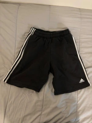 Svarta Adidas shorts med vita ränder - Klassiska svarta Adidas shorts med vita ränder längs sidorna och Adidas-logga på vänster ben. Elastisk midja och bekväm passform, perfekta för träning eller chill. Tillverkade i polyester för en sportig känsla.
