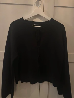 Svart stickad tröja Gina Tricot - Svart stickad tröja från Gina Tricot med långa ärmar och knytning framtill. Tröjan har en enkel, rak passform och rund halsringning. Perfekt att styla med jeans eller kjol för en clean look.