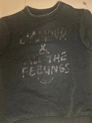 Mörkgrå sweatshirt med texttryck - Mysig mörkgrå sweatshirt med glittrigt texttryck 'L'AMOUR & ALL THE FEELINGS' på bröstet. Tröjan har rund halsringning, långa ärmar och ribbade muddar vid ärmslut och nederkant. Perfekt för en avslappnad och trendig look.