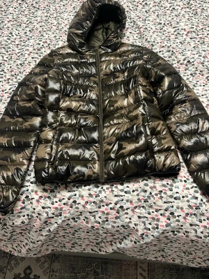 Kamo mönstrad dunjacka med huva - Snygg dunjacka med glansig camouflage-mönster i gröna och svarta toner. Jackan har huva, dragkedja framtill och quiltad design. Perfekt för kalla dagar när du vill ha både stil och värme.