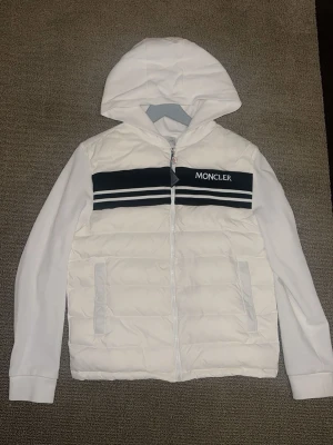 Vit Moncler jacka med huva - Snygg vit jacka från Moncler med huva och dragkedja. Jackan har vadderad front, två fickor och svarta samt vita ränder över bröstet med Moncler-logga. Ärmar och rygg är i mjukt tyg medan framsidan är quiltad. Perfekt för dig som vill ha en stilren och sportig look.