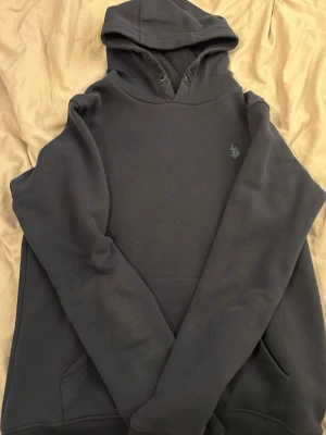 Mörkblå hoodie från Polo Assn - Snygg mörkblå hoodie från Polo Assn med klassisk huva och känguruficka. Diskret broderad logga på bröstet i vitt. Tillverkad i mjukt bomullsmaterial som känns skönt mot huden. Perfekt för en avslappnad och stilren look.