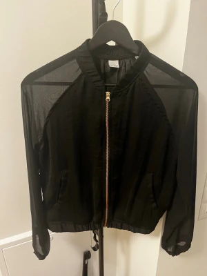 Svart mesh vindjacka med dragkedja - Snygg svart vindjacka med transparenta meshärmar och axelparti. Jackan har dragkedja framtill, två fickor och resår i midjan. Perfekt för en trendig streetstyle-look. Materialet är lätt och luftig, vilket gör den enkel att bära över en t-shirt.