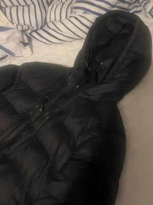 Zavetti canada jacka  - Säljer en svart dunjacka från zavetti canada med huva och dragkedja framtill. Jackan har flera fickor med dragkedja och en puffig, quiltad design som håller dig varm under kalla dagar. Perfekt för dig som vill ha en stilren och funktionell vinterjacka.