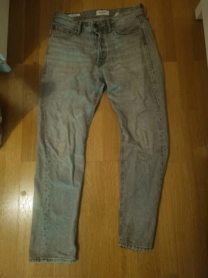 Jack & jones, ljusgråa jeans  - Säljer ett par ljusgrå jeans med rak passform och klassisk femficksdesign. Jeansen har en enkel look utan slitningar eller dekorationer, och är tillverkade i ett mjukt denimtyg. Perfekta för en avslappnad stil.