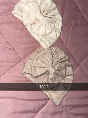 Två turbanmössor i beige och offwhite - Två mjuka turbanmössor i stickad look, en i beige och en i offwhite. Båda har ett dekorativt knut framtill och diskret mönster i tyget. Perfekt accessoar för att lyfta din stil.