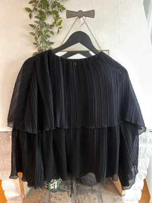 Plisserad svart blus med volanger - Snygg svart blus med plisserat tyg och dubbla lager volanger som ger ett luftigt och trendigt intryck. Blusen har rund halsringning och vida, långa ärmar. Perfekt för dig som gillar en elegant och modern stil.