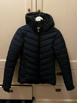 Mörkblå skidjacka - Snygg mörkblå skidjacka med huva och dragkedja framtill. Jackan är från Mckinley och är ett par år gammal, men funkar fint och är hel, förutom en söm som gått upp vid ena mudden. Den har en liten ficka för liftkort och även snölås. Perfekt jacka för kallare dagar! Säljer då jag köpt en annan jacka och inte behöver den här längre ☺️ vid snabb affär kan priset gå att diskutera :)