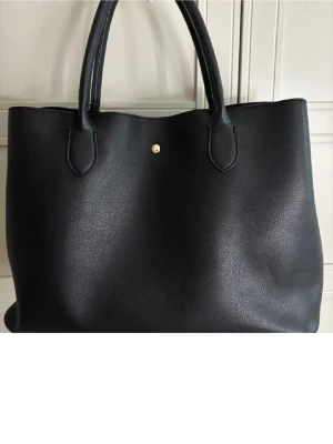 Svart tote handväska i konstläder - Stilren svart tote handväska i konstläder med två rejäla handtag och guldfärgad knappdetalj. Väskan har ett rymligt huvudfack och en innerficka med dragkedja för extra förvaring. Perfekt för dig som vill ha plats för allt du behöver under dagen.