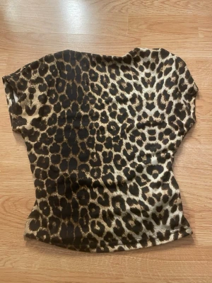 Leo T-shirt med öppenrygg  - Säljer en trendig t-shirt med leopardmönster i bruna och svarta toner. T-shirten har korta ärmar och en figurnära passform. Perfekt för dig som vill sticka ut med ett djurmönster. Materialet känns mjukt och stretchigt, troligen polyester eller bomull.