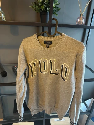 Grå stickad tröja från Polo Ralph Lauren - Snygg grå stickad tröja från Polo Ralph Lauren med stor POLO-text i svart och vitt framtill. Tröjan har rund halsringning, ribbade muddar och är perfekt för dig som gillar klassisk streetwear med en sportig touch.