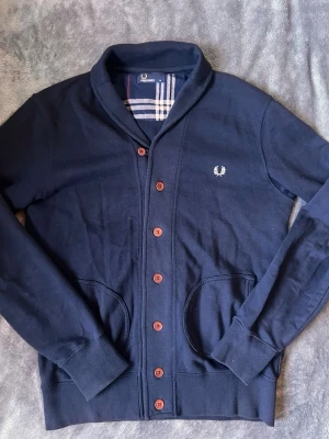 Mörkblå cardigan från Fred Perry - Snygg mörkblå cardigan från Fred Perry med bruna knappar och klassisk logga broderad på bröstet. Tröjan har två fickor framtill, ribbade muddar och en mjuk krage. Tillverkad i bomull för en bekväm känsla och stilren look.