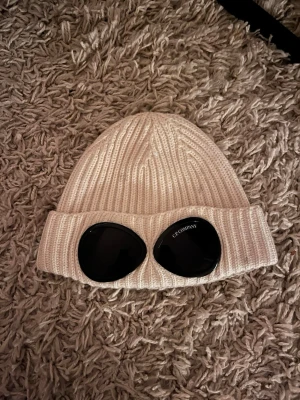 Vit mössa C.P. Company - Säljer en vitmössa från C.P. Company med ikoniska svarta goggles framtill. Mössan har uppvikt kant och är tillverkad i mjukt, stretchigt material som sitter skönt på huvudet. Perfekt accessoar för en trendig streetwear-look.