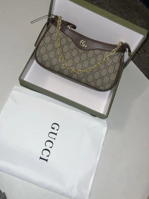 Gucci GG Supreme axelremsväska brun - Snygg axelremsväska från Gucci i klassiskt GG Supreme-mönster med bruna läderdetaljer och guldfärgad kedja. Väskan har en elegant, avlång form och logga framtill. Perfekt för dig som vill ha en ikonisk och trendig accessoar.