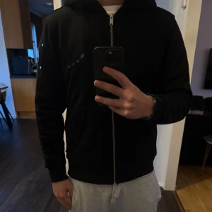 Äkta Svart Moose Knuckles  jacka - Svart hoodie från Moose Knuckles med silverfärgad dragkedja och klassisk logga i metall på ärmen. Tröjan har huva, känguruficka och är långärmad. Perfekt för dig som gillar streetwear och vill ha en stilren look.