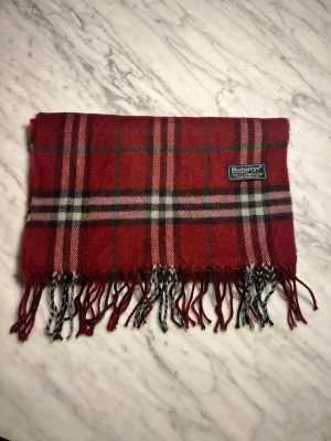 Röd halsduk från Burberry - Klassisk röd halsduk från Burberry i ull med rutigt mönster i svart, vitt och grönt. Halsduken har fransar i ändarna och en vävd logotypetikett. Perfekt accessoar för att lyfta din outfit.