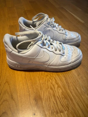 Nike Air Force 1 vita sneakers - Klassiska vita Nike Air Force 1 sneakers i lågt utförande. Skorna har en stilren design med perforerad tå, vita skosnören och den ikoniska swoosh-loggan på sidan. Tillverkade i läder med en robust gummisula och AIR-dämpning för extra komfort.