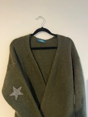 Olivgrön kofta med stjärndetalj - Mysig olivgrön kofta från Funky Staff med djup V-ringning och lång ärm. På vänster ärm finns en glittrig silverfärgad stjärna som snygg detalj. Koftan är stickad i ett mjukt material och har en avslappnad passform.