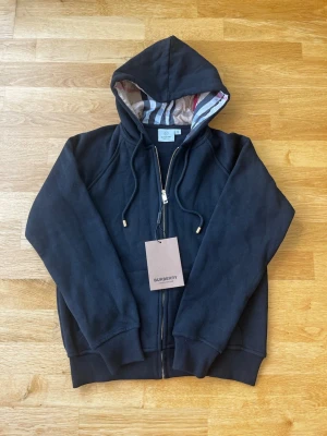 Svart hoodie från Burberry - Snygg svart hoodie från Burberry med dragkedja och huva. Insidan av huvan har det klassiska Burberry-mönstret i beige, rött och blått. Hoodien har ribbade muddar och fickor framtill. Perfekt för dig som vill ha en stilren och exklusiv look. Den är helt ny aldrig använt och priset kan diskuteras.
