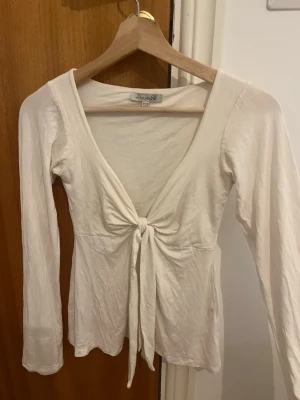Vit långärmad topp med knyt - Supersnygg vit långärmad topp från Designers Society med djup v-ringning och knyt framtill. Toppens material känns mjukt och lätt, perfekt för att skapa en chill och trendig look. Passar perfekt till jeans eller kjol för en stilren outfit.