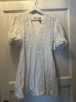 Vit studentklänning  - Kort vit klänning från Zara med puffärmar och v-ringning. Klänningen har ett broderat mönster över hela tyget. Jättefin studentklänning som aldrig har använts endast tvättad 1 gång. 