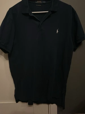 Mörkblå pikétröja från Ralph Lauren - Klassisk mörkblå pikétröja från Ralph Lauren med vit broderad logga på bröstet. Tröjan är i Pima stretch mesh-material och har korta ärmar samt krage med knappslå. Perfekt för en stilren och avslappnad look.