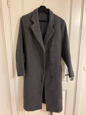 Grå ullrock från Filippa K - Filippa K kappa för dam i storlek S. Lång modell i grå ull med klassisk krage och knäppning fram. Avtagbart knytskärp i midjan. Fodrad insida för ökad komfort. Passar som ytterplagg under höst och vinter. Den är jätte fin och jätte bra skick. Det står medium i storleken, men den är S/36. Materilet är ull 75%.