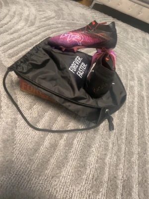 Puma lila och svarta fotbollsskor - Säljer ett par snygga Puma fotbollsskor i lila och svart med orange detaljer. Skorna har låg vikt, snörning och en modern design med mönstrad ovandel. Kommer med en svart Puma-påse med texten 'FOREVER. FASTER.' Perfekta för dig som vill sticka ut på planen. Och dom är aldrig använda