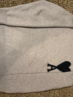 Grå stickad tröja från Ami - Snygg grå stickad tröja från Ami Alexandre Mattiussi med svart hjärtdetalj och bokstaven A på bröstet. Tröjan har ribbade muddar vid ärmslut och nederkant samt en klassisk rund hals. Perfekt för dig som gillar stilrena plagg med en twist. Storlek S men kan räknas som M.