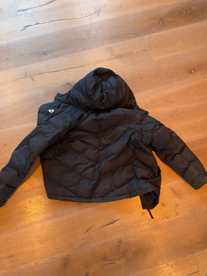 Svart pufferjacka från Nike XL barn  - Svart pufferjacka från Nike med huva och dragkedja. Jackan har en klassisk quiltad design och är perfekt för kalla dagar. Märket syns tydligt på ärmen och insidan. Passar dig som gillar sportig och stilren look. Storlek 158-170
