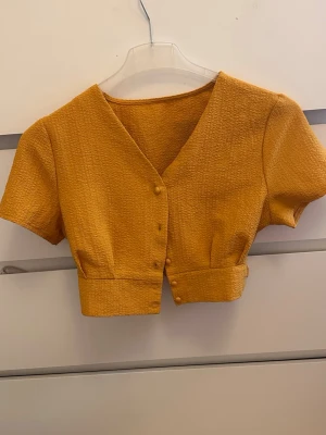 Gul croppad topp med knappar - Trendig gul croppad topp med v-ringning och korta ärmar. Toppen har dekorativa knappar framtill och är gjord i ett strukturerat tyg som ger en cool look. Perfekt att styla med höga jeans eller kjol.