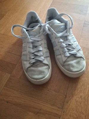 ljusblåa Adidas sneakers med ränder - Klassiska vita Adidas sneakers med de ikoniska tre ränderna på sidan. Skorna har lågt skaft, snörning och ovandel i mocka och textil. Perfekta för en clean och avslappnad stil. Diskret Adidas-logga på plösen.