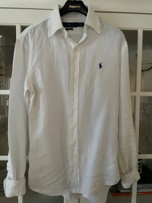 Vit linneskjorta från Ralph Lauren - Klassisk vit skjorta från Ralph Lauren i linne med blå broderad logga på bröstet. Slim fit-modell med långa ärmar och knappar framtill. Perfekt för en clean och stilren look.